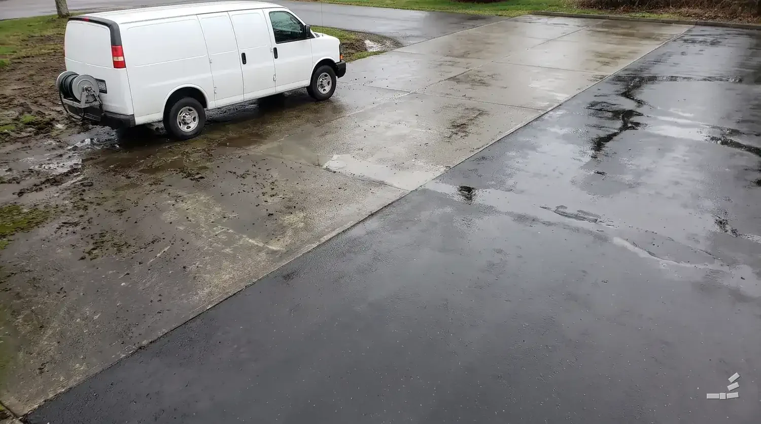 Photograph-style white work van at the edge of an asphalt lot, stained asphalt on the near side of the diagonal and wet clean asphalt beyond it