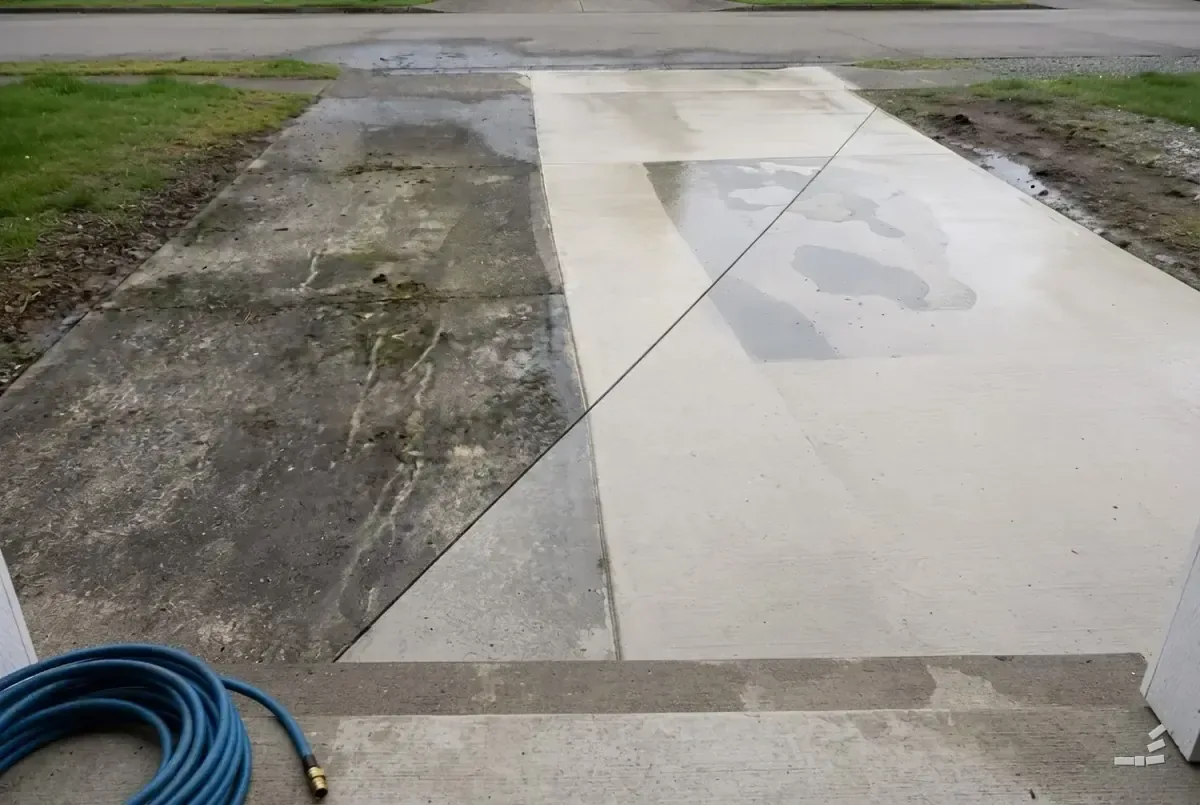 Photograph-style residential front walkway with one cleaned pass running down the middle of a stained concrete drive, a coiled hose in the foreground