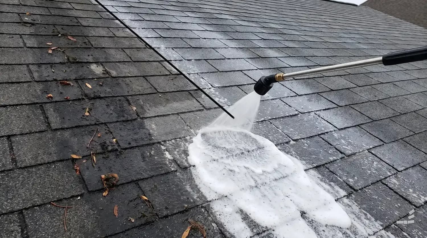 Photograph-style asphalt shingle roof being soft washed with low-pressure foam, half still algae-streaked
