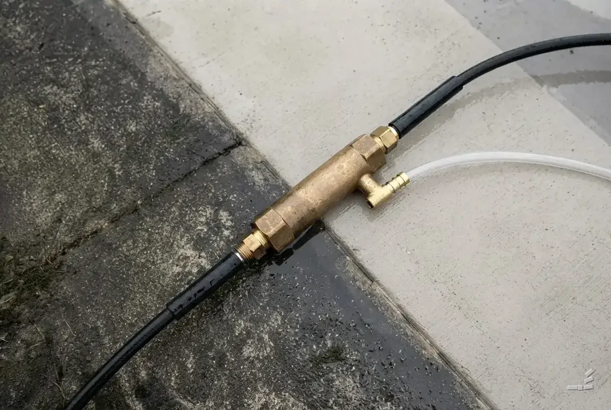 Photograph-style brass downstream injector on a concrete slab, a black hose screwed into each end and a clear chemical tube pushed onto its side barb