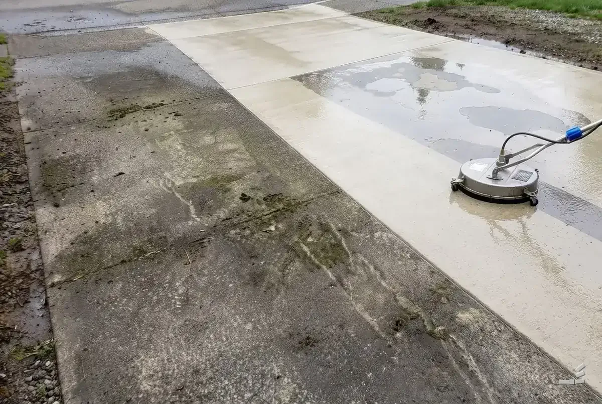 Photograph-style surface cleaner working a driveway apron, mossy stained concrete on one side of the cleaned edge and bare wet clean concrete on the other